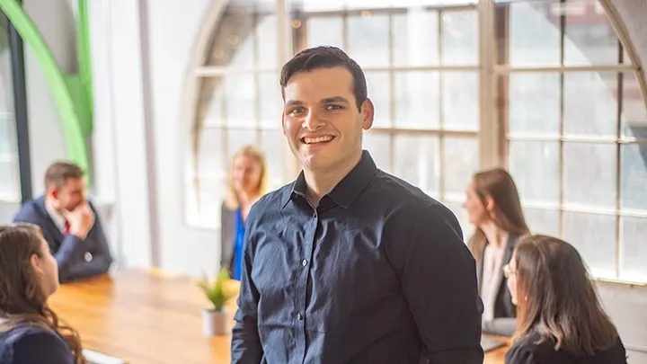 Man smiling at the camera while a meeting takes place behind him.