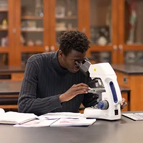 Male college student sitting at lab table looking through microscope